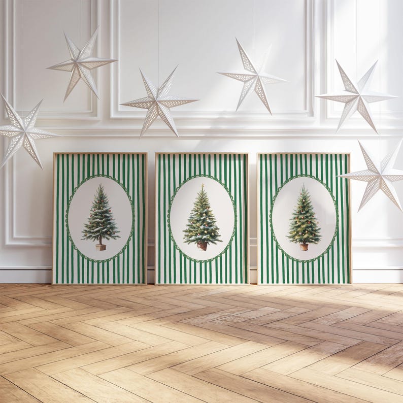 May include: Three framed Christmas tree prints with green and white striped backgrounds. Each print features a watercolor-style Christmas tree in an oval frame. White star-shaped decorations hang from the ceiling. The floor is made of wood.