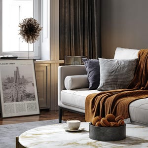 K&ouml;nnte beinhalten: Ein Wohnzimmer mit einem wei&szlig;en Sofa, Kissen und einer rostfarbenen Decke. Ein Marmor-Couchtisch zeigt eine Schale mit Keksen und eine Kaffeetasse. Ein gerahmter Druck mit Text ist in der N&auml;he eines Fensters zu sehen.