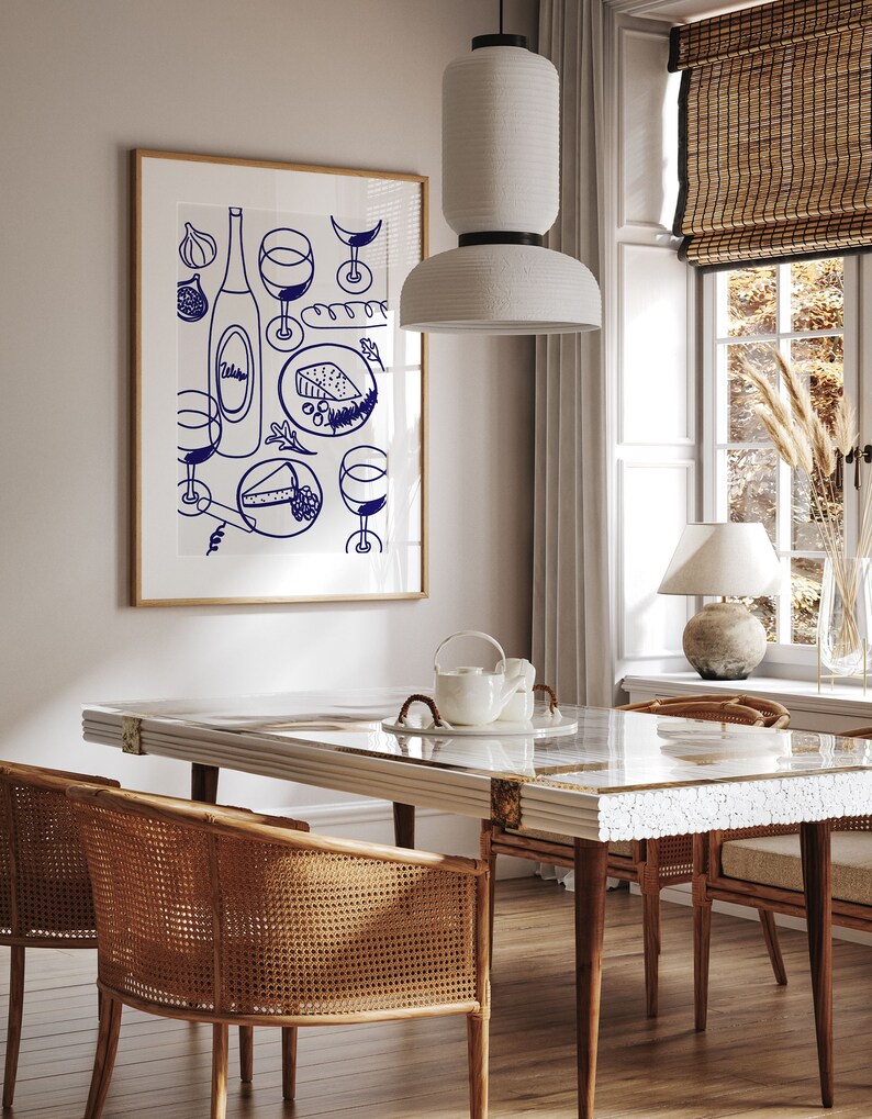 May include: A white dining table with a white marble top and a white ceramic teapot and cups on a white plate. Two wicker chairs are on either side of the table. A framed print of a blue and white illustration of wine glasses, a bottle of wine, cheese, and grapes hangs on the wall behind the table.