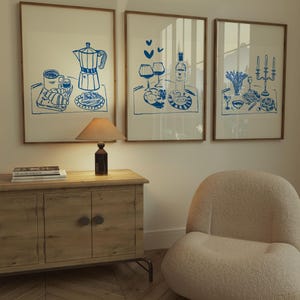 May include: Three framed illustrations of breakfast, lunch, and dinner scenes. Each illustration is in blue ink on a white background. The breakfast scene shows a coffee pot, a cup of coffee, a croissant, and a slice of toast. The lunch scene shows two glasses of wine, a bottle of wine, and a plate of food. The dinner scene shows a plate of food, a vase of flowers, and two candlesticks.