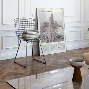 K&ouml;nnte beinhalten: Ein modernes Wohnzimmer mit einem Drahtstuhl mit B&uuml;chern, einem gerahmten Poster mit dem Text "F1 Saudi Arabia" und einer kleinen Schale auf einem Holztisch. Der Raum hat wei&szlig;e W&auml;nde und Parkettboden.