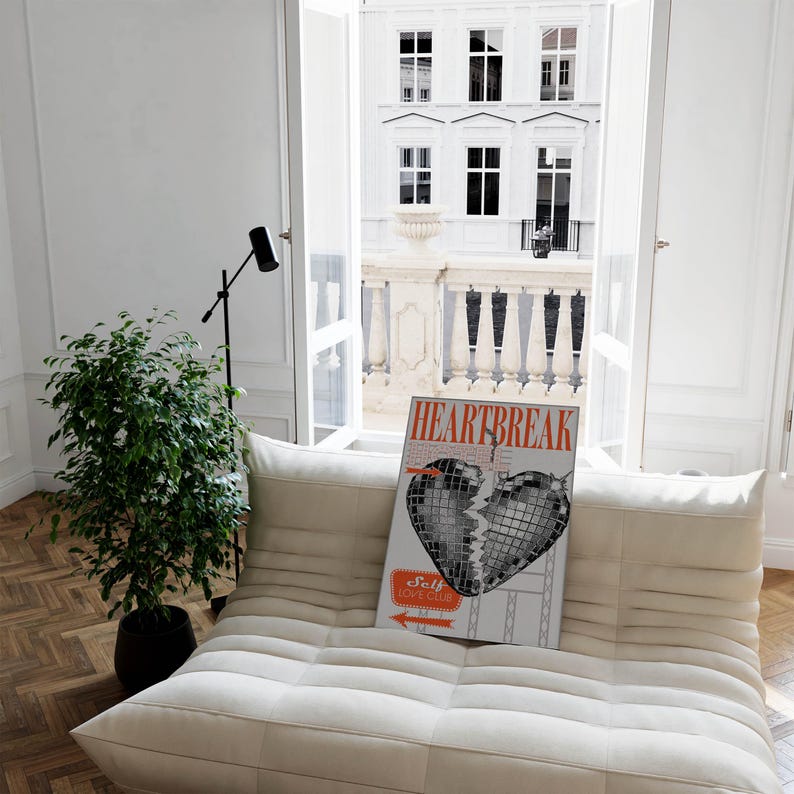 Pode incluir: Um sof&aacute; branco com um p&ocirc;ster em preto e branco de um cora&ccedil;&atilde;o partido com o texto "HEARTBREAK" e "Self Love Club". O p&ocirc;ster est&aacute; em um sof&aacute; branco em uma sala com uma janela e uma planta.