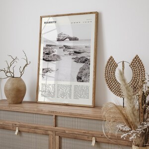 Pu&ograve; includere: Una fotografia in bianco e nero incorniciata di Biarritz, Francia, con una scena costiera con rocce e una formazione rocciosa ad arco. La cornice in legno &egrave; su un mobile in legno con un vaso ed elementi decorativi.