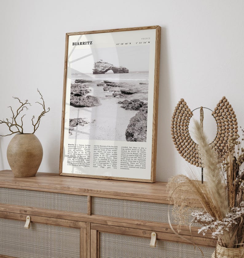 Pu&ograve; includere: Una fotografia in bianco e nero incorniciata di Biarritz, Francia, con una scena costiera con rocce e una formazione rocciosa ad arco. La cornice in legno &egrave; su un mobile in legno con un vaso ed elementi decorativi.