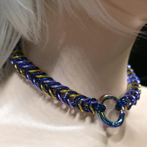 May include: A multi-colored chainmail necklace with blue, gold, and purple rings. The necklace has a large, iridescent O-ring clasp. The chainmail is woven in a tight pattern, creating a textured appearance.