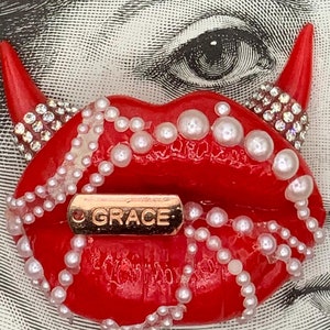 May include: Red lips with a gold 'GRACE' tag and white pearl accents. The lips are part of a larger design with devil horns.