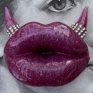 May include: A pair of purple lips with glitter, shaped like a devil's horns with rhinestones.