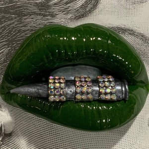 May include: A pair of green lips with a silver bullet with rhinestones in the middle of the lips.