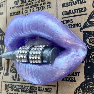 May include: A purple, glittery, silicone mold of a pair of lips biting down on a silver and black bullet with rhinestones. The mold is sitting on a brown surface with text.
