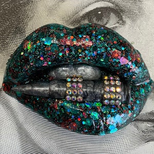 May include: A pair of black lips with green, red, and blue glitter are holding a silver bullet with rhinestones.