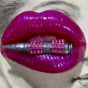 May include: A close-up of a pair of bright pink lips with a silver bullet covered in pink rhinestones in the middle.