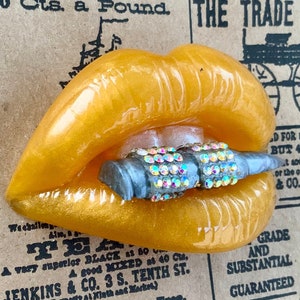 May include: A golden-yellow, glossy lips-shaped object with a silver-colored object adorned with iridescent rhinestones. The lips are set against a vintage newspaper background.