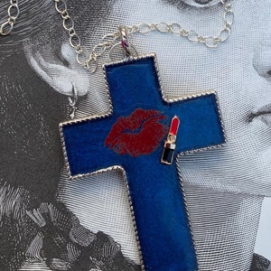 May include: A silver chain necklace with a blue cross pendant. The cross has a red lip print and a gold lipstick design on it.