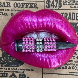 May include: A pink, glossy, sculpted pair of lips with a black and silver bullet with pink rhinestones in the mouth. The lips are on a brown background with text.