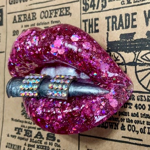 May include: Pink glitter resin lips with a silver bullet charm. The lips are set against a vintage newspaper background with text that reads "The Trade Wagon".