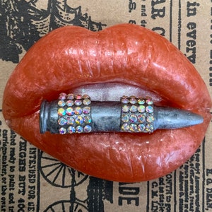 May include: A resin sculpture of coral-colored lips with a silver bullet held between two rhinestone-studded bars. The background features vintage newspaper print with text including "UNDERWEAR" and "BIG MONEY!"