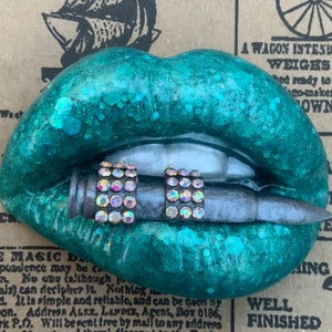 May include: A teal green resin lips sculpture with a silver bullet in the mouth. The bullet is decorated with iridescent rhinestones.