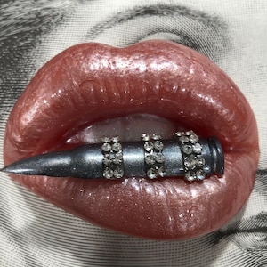 May include: A close-up of a pair of red lips with a silver bullet adorned with rhinestones in the center.