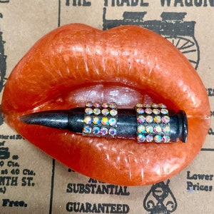 May include: A resin art piece featuring a pair of orange lips holding a black bullet. The bullet is adorned with iridescent rhinestones. The background is a collage of vintage newspaper clippings.