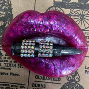 May include: A pink resin sculpture of a pair of lips with a black bullet with rhinestones in between the lips. The sculpture is on a vintage newspaper background.
