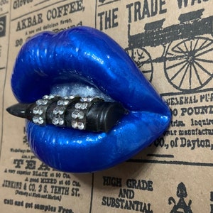 May include: A blue, glossy, sculpted pair of lips with a black and silver studded bullet piercing through the center. The lips are set against a vintage newspaper background with text that reads "The Trade Wagon."