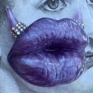May include: A pair of purple lips with a sparkly finish, each lip has a small, pointed horn with rhinestones.