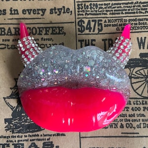 May include: A pink resin lip charm with silver glitter and rhinestones. The charm has two pink devil horns with rhinestones. The charm is on a brown background with text.