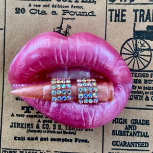 May include: Pink lips with a bullet in between them. The bullet is decorated with rhinestones. The background is a vintage newspaper.