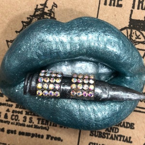 May include: Teal blue lips with a black bullet with rhinestones. The bullet is in the mouth of the lips.