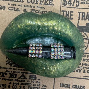 May include: Green and gold glitter lips with a black bullet with rhinestones embedded in the sides. The lips are open and the bullet is in the mouth.