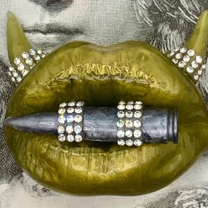 May include: A pair of gold lips with devil horns and rhinestones, holding a silver bullet with rhinestones.