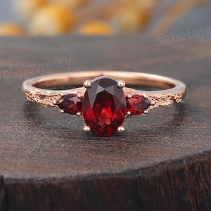 May include: Rose gold ring featuring an oval-cut, deep red gemstone flanked by two smaller gemstones. The band has a textured, branch-like design. A piece of jewelry.