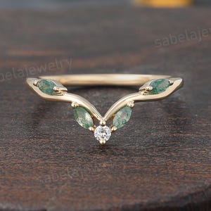 May include: A gold ring with a curved design. The ring features three green marquise-cut gemstones and a small, round, clear gemstone. The ring is set against a dark brown wooden surface.