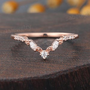 Peut inclure: Bague en or rose avec un design en forme de V. La bague présente un petit diamant rond au centre et des diamants taille marquise et ronds le long de la bande. La bague est posée sur une surface en bois foncé.