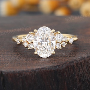 Pode incluir: Um anel de ouro com um grande diamante central de corte oval, ladeado por grupos de diamantes menores. O anel está sobre uma superfície de madeira escura. O design é elegante, adequado para noivados ou ocasiões especiais.