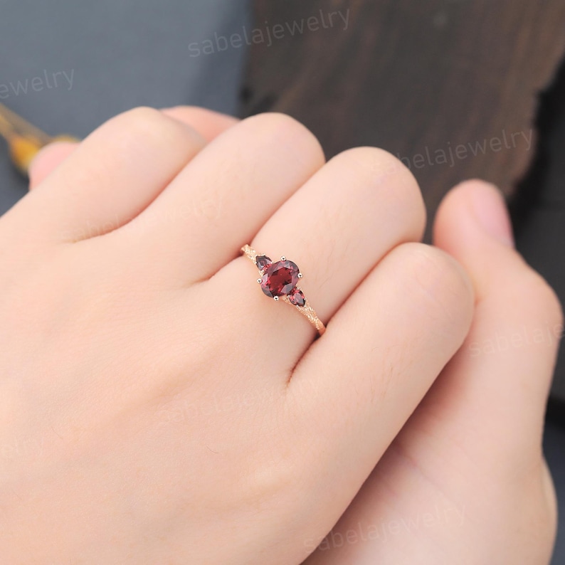 May include: A rose gold ring featuring a central oval-cut garnet gemstone, flanked by two smaller garnet stones. The ring's band has a textured design, and the ring is displayed on a hand. The text "sabelajewelry" is visible in the background.