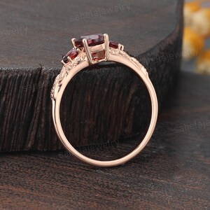 May include: Rose gold ring featuring three oval-cut, deep red gemstones. The ring has an ornate, floral-inspired design on the band. The ring is displayed on a dark wooden surface, showcasing the jewelry's intricate details.