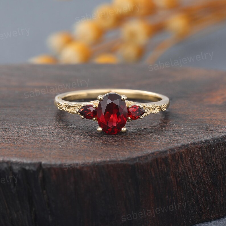 May include: A gold ring featuring a large oval-cut, deep red gemstone flanked by two smaller red gemstones. The band has an ornate, textured design. The ring is displayed on a dark wooden surface.