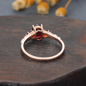 May include: Rose gold ring featuring an oval-cut, dark red gemstone as the centerpiece, flanked by smaller red gemstones. The band has a delicate, ornate design. The ring is set against a dark wooden surface.