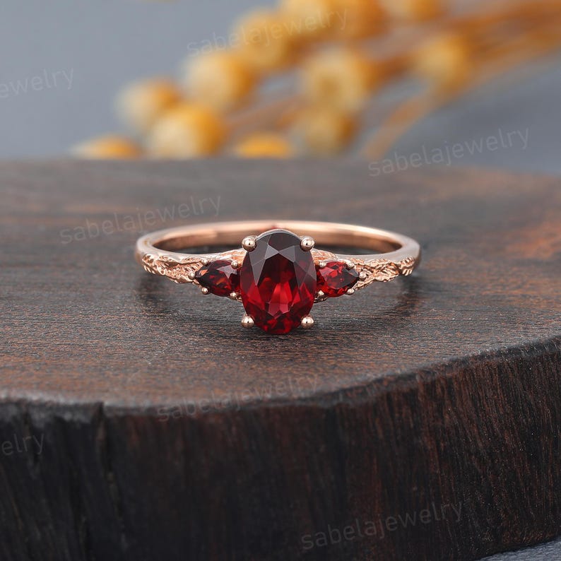 May include: Rose gold ring featuring an oval-cut, deep red gemstone flanked by two smaller gemstones. The band has a textured, branch-like design. A piece of jewelry.