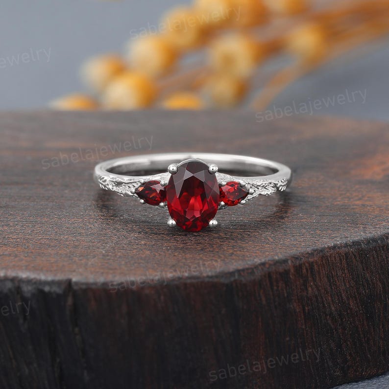 May include: A silver ring with an oval, deep red gemstone at the center, flanked by two smaller red gemstones. The band has a textured design. The ring is displayed on a dark wooden surface.