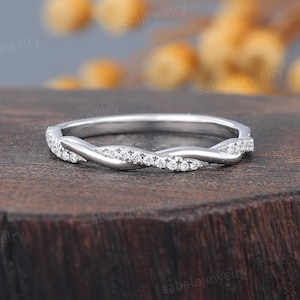 May include: A silver ring with a twisted design, set with small, clear gemstones. The ring is displayed on a dark wooden surface. The gemstones add a subtle sparkle to the ring's design.
