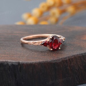 May include: Rose gold ring featuring an oval-cut, deep red gemstone as the centerpiece, flanked by two smaller triangular gems. The band has an intricate, textured design. The ring is displayed on a dark wooden surface, showcasing its elegant details.