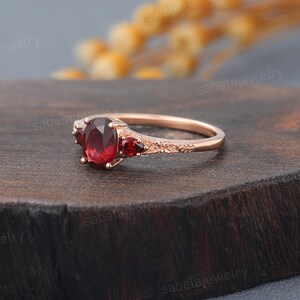 May include: Rose gold ring featuring a large oval-cut red gemstone flanked by two smaller red gemstones. The band has an intricate, textured design. The ring is displayed on a dark wooden surface.