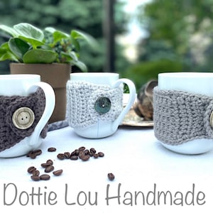 Op de afbeelding: Drie witte keramische mokken met gehaakte grijze en bruine mokwarmers. Elke warmer heeft een knoopsluiting. De mokken staan op een wit oppervlak met koffiebonen die eromheen zijn verspreid. De tekst "Dottie Lou Handmade" staat onderaan de afbeelding.