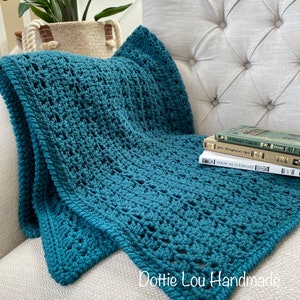 May include: A teal blue crocheted throw blanket draped over a white armchair with a stack of books on the seat. The blanket has a lacy, open stitch pattern. The text "Dottie Lou Handmade" is visible in the bottom right corner of the image.