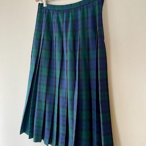 Puede incluir: Una falda plisada con estampado de cuadros verdes y azules cuelga de una percha de madera. La falda tiene un diseño clásico con pliegues verticales y cierre de botón. La tela presenta un patrón de cuadros verde oscuro, azul marino y negro.