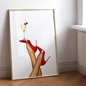May include: A framed art print featuring a pair of legs wearing red high heels, supporting a champagne glass with cherries. The artwork has a gold frame and is leaning against a white wall, with a window and radiator in the background.
