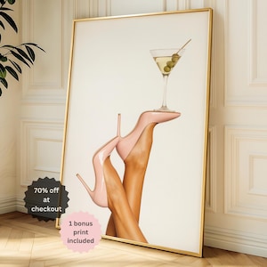 May include: A framed art print featuring a martini glass balanced on a pair of pink high-heeled shoes. The martini contains olives. The print is in a gold frame and leans against a white wall. The image includes the text "70% off at checkout" and "1 bonus print included".