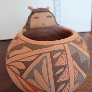 Könnte beinhalten: Ein kleiner, runder Terrakotta-Topf mit einem geometrischen Muster in Braun-, Rot-, Blau- und Weißtönen. Der Topf hat oben eine kleine, geformte Figur einer Person mit braunem Kopf und weißen Augen.
